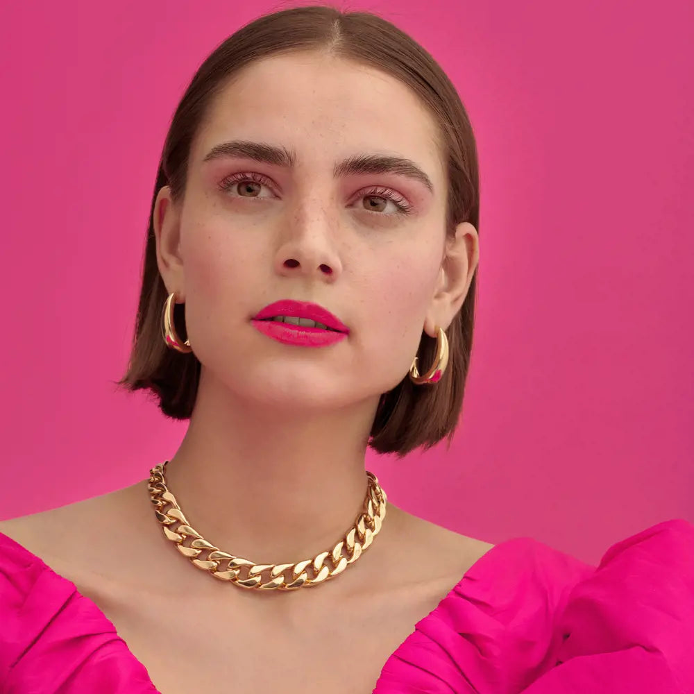 Woman wearing a gold necklace and hoop earrings against a pink background