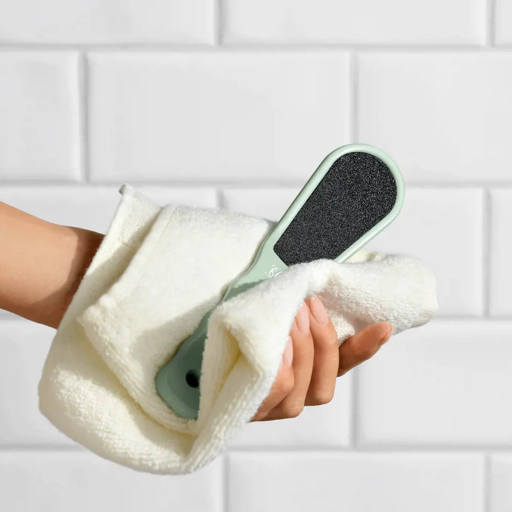 Hand holding a green foot scrubber with a towel against a tiled wall background
