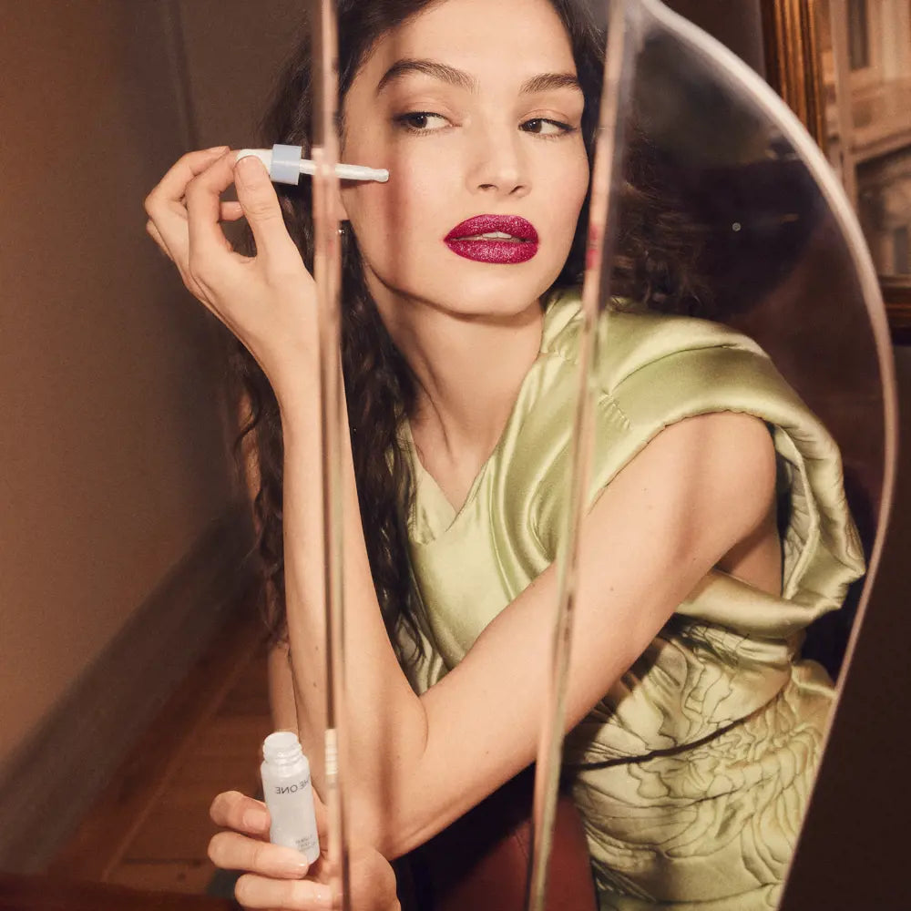 Woman applying makeup with a brush and bottle, wearing a green dress.